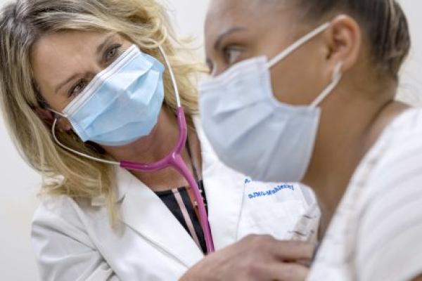 A doctor holds her stethoscope up to a patient’s upper chest, listening to her heartbeat. Both the physician and the patient are wearing surgical masks.